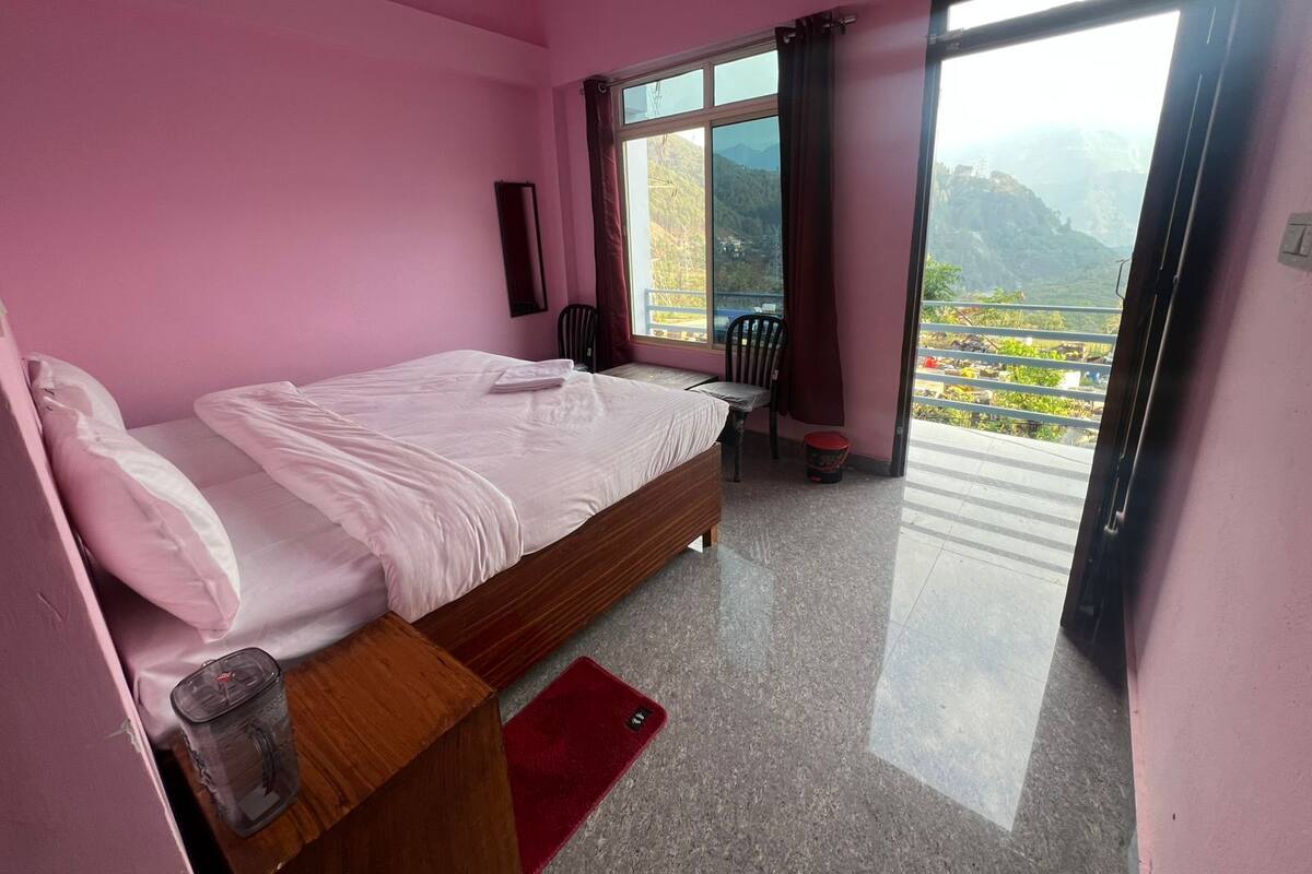 A pink-themed bedroom with a bed, a chair, a small table, and a view of a mountainous landscape through a large window.