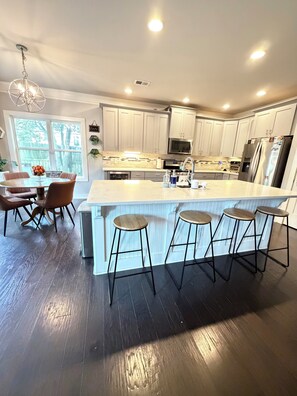 A modern kitchen with white cabinets, a central island with stools, a dining area with a table and chairs, and a large window with a view of trees. - Amazing 4 Bedroom Home Away from Home for World Cup/ Atlanta Vacation (Smyrna)