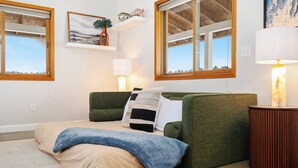 A bedroom with a bed, a green sofa, a wooden side table, a lamp, and two windows. - Northern Escape - Hot Tub, Sauna, Cold Plunge (Lake Ann)