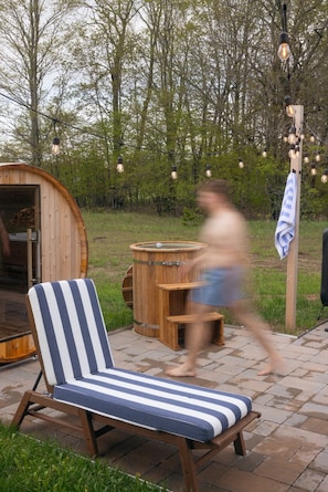A wooden barrel sauna, a striped lounge chair, and a wooden bench. - Northern Escape - Hot Tub, Sauna, Cold Plunge (Lake Ann)