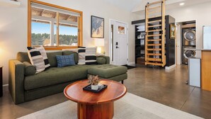 A living room with a green sofa, a wooden coffee table, a large window, and a sliding door leading to a laundry area. - Northern Escape - Hot Tub, Sauna, Cold Plunge (Lake Ann)