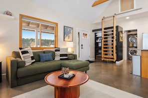 A living room with a green sectional sofa, a wooden coffee table, a large window, and a sliding closet door. - Northern Escape - Hot Tub, Sauna, Cold Plunge (Lake Ann)