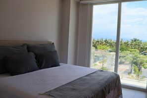 Ein Bett mit weißer und grauer Bettdecke, einem Kopfteil und Blick auf Palmen durch ein großes Fenster.