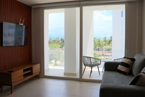 Ein modernes Wohnzimmer mit Flachbildfernseher, einer Holz-Unterhaltungseinheit, einem grauen Sofa mit gestreiften Kissen und einer Glasschiebetür, die auf einen Balkon mit Blick auf Palmen und Gebäude führt.