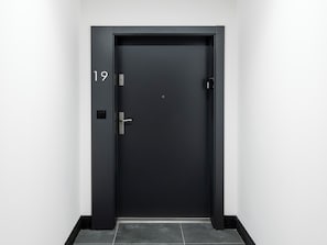 Eine schwarze Tür mit Zahlenschloss und Griff, eingebaut in einen Flur mit weißen Wänden und dunklen Fliesen.