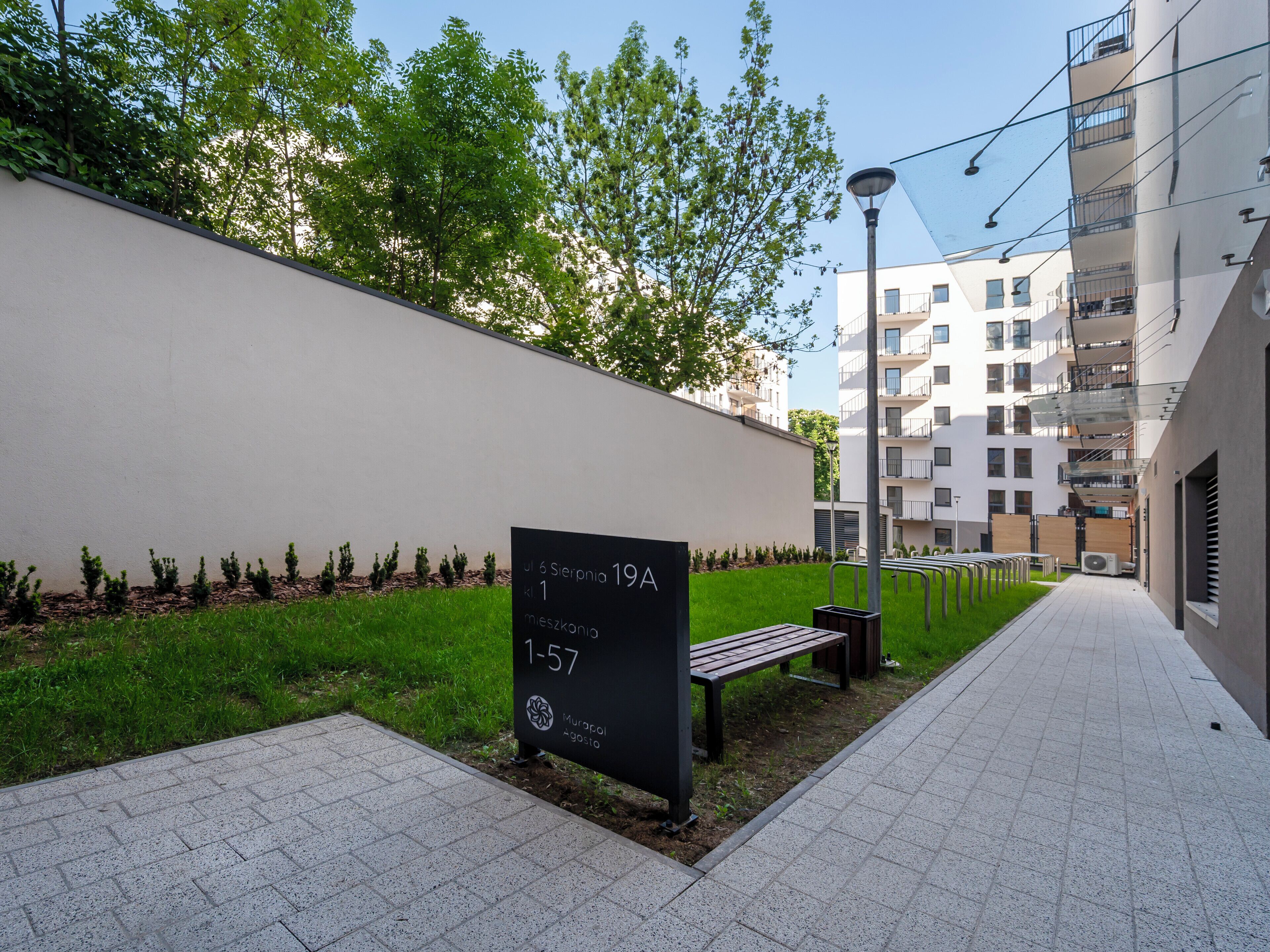 Un camino pavimentado, un banco, un cartel con números de apartamentos y un edificio con balcones.