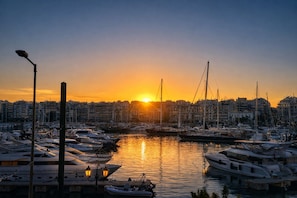 Un port de plaisance avec des bateaux amarrés, un coucher de soleil en arrière-plan et des bâtiments le long de la promenade.