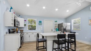 A modern kitchen with white cabinetry, a central island with bar stools, a built-in oven, and a window with blinds. - Sunset Drift Cottage (Fernandina Beach)
