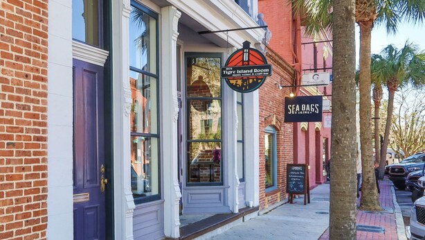 A brick building with a shop entrance, a sign for 'Tig Island Room' and 'Sea Bags,' and a sidewalk with palm trees. - Sunset Drift Cottage (Fernandina Beach)