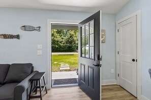 Eine offene Tür führt zu einer grünen Wiese, eine graue Couch und ein kleiner Tisch sind zu sehen.