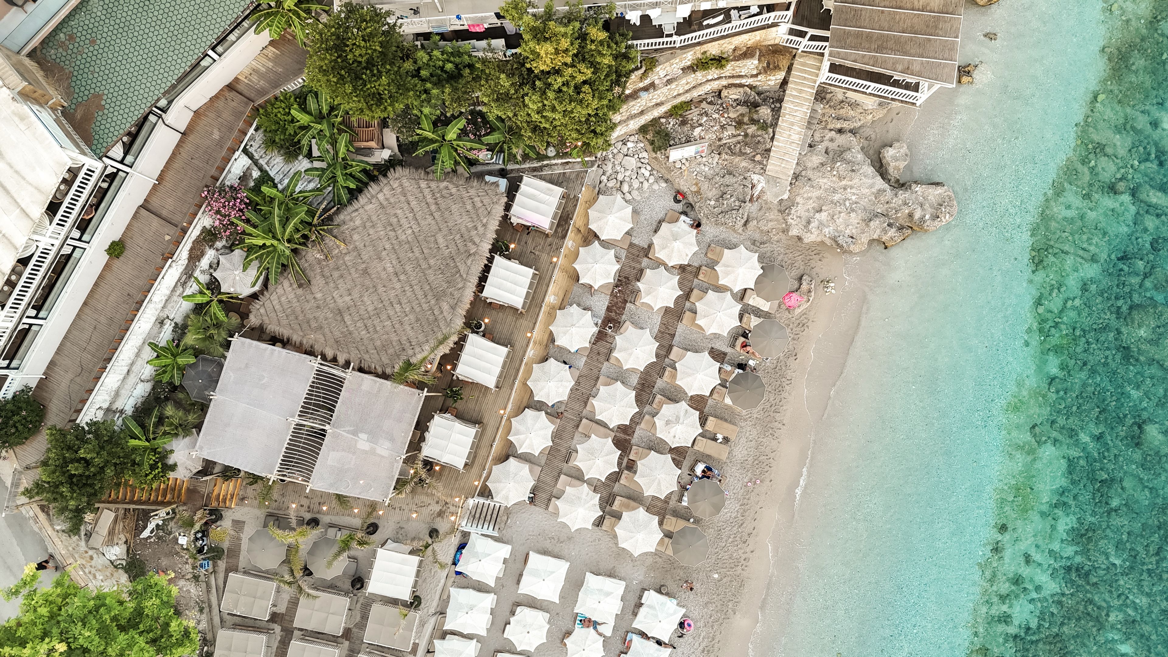 Vista aérea de un hotel frente a la playa con varias zonas de estar cubiertas por sombrillas y una estructura con techo de paja.