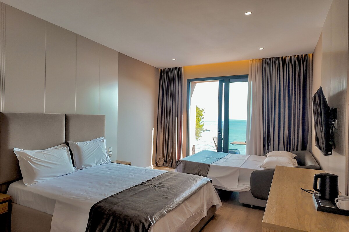 A hotel room with a bed, a desk, a chair, a TV, and a view of the sea.