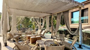 A covered outdoor seating area with wicker furniture and a pool in the background. - Hotel Platinium Dhermi (Dhermi)