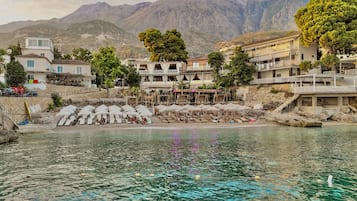 A coastal hotel with multiple balconies, a pool area, and a view of mountains in the background.