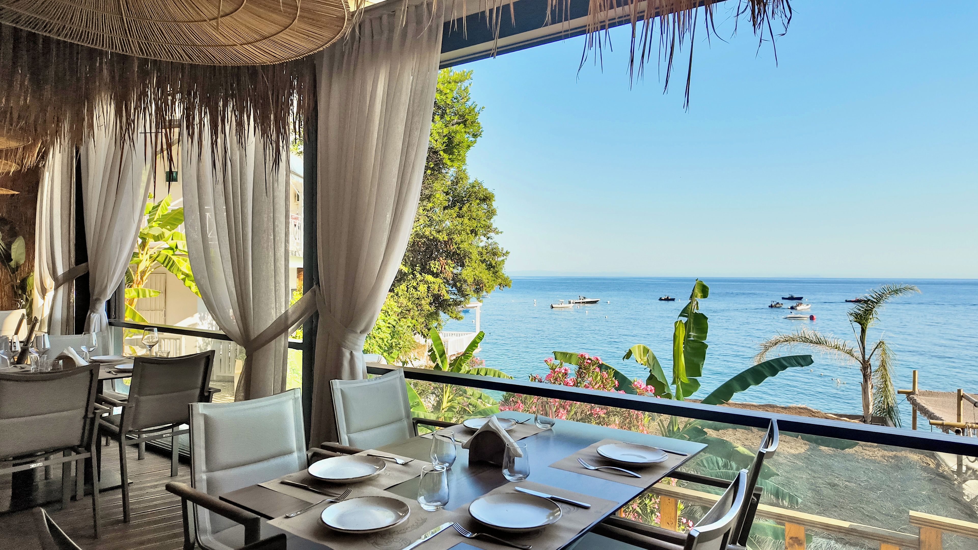 Comedor con una mesa lista para dos personas, con vista al mar azul y algunas embarcaciones.
