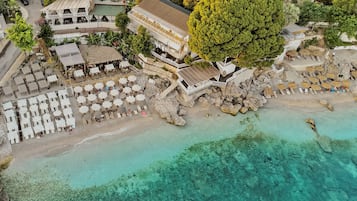 An aerial view of a beachside resort with multiple lounge areas, a large tree, and clear turquoise waters.