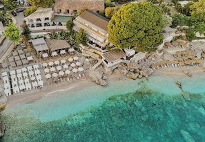 Vista aérea de un complejo turístico frente a la playa con varias zonas de descanso, un árbol grande y aguas turquesas cristalinas.