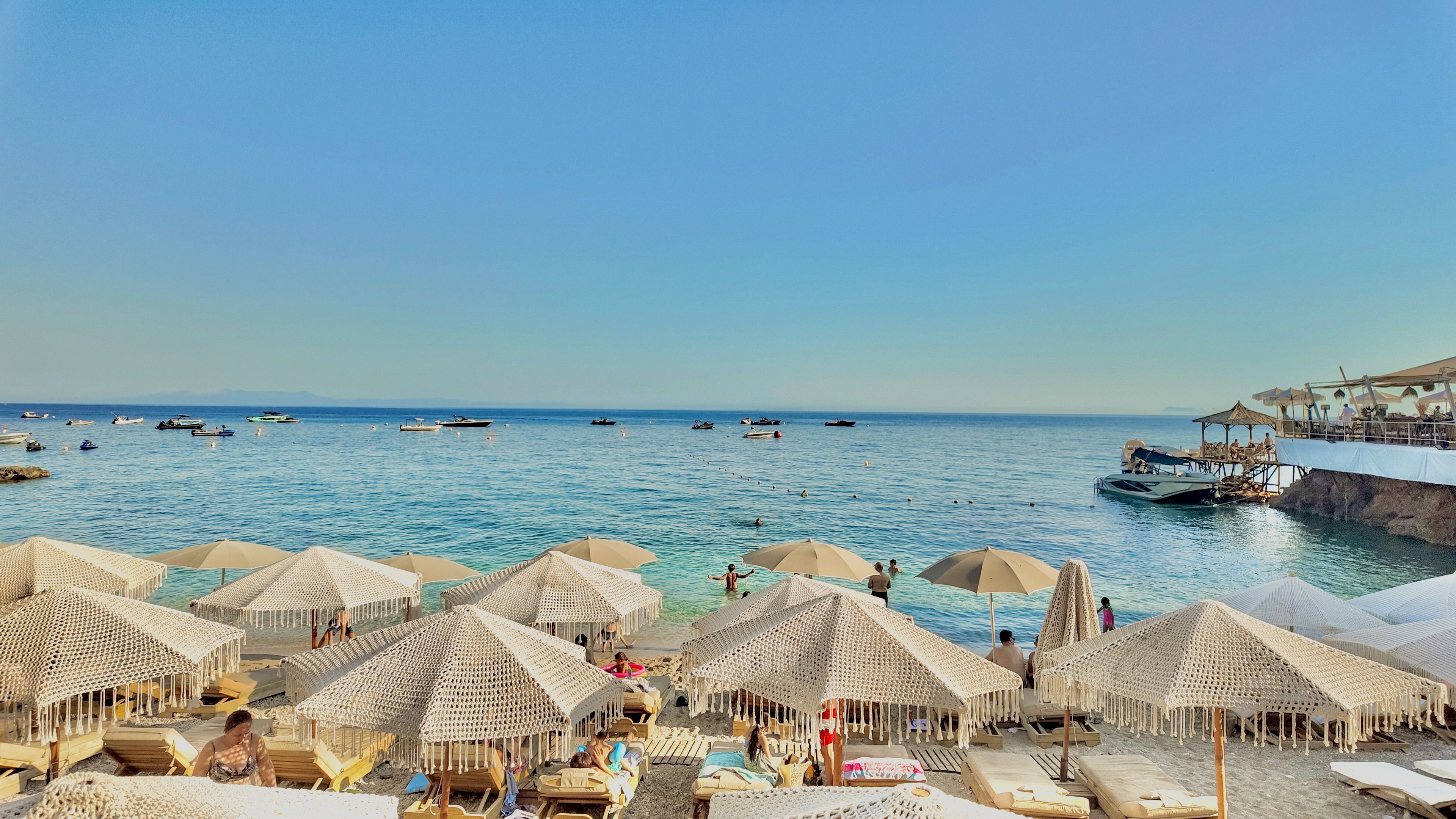 Una playa con sombrillas de paja y sillas reclinables, barcos en el agua y un muelle con restaurante.