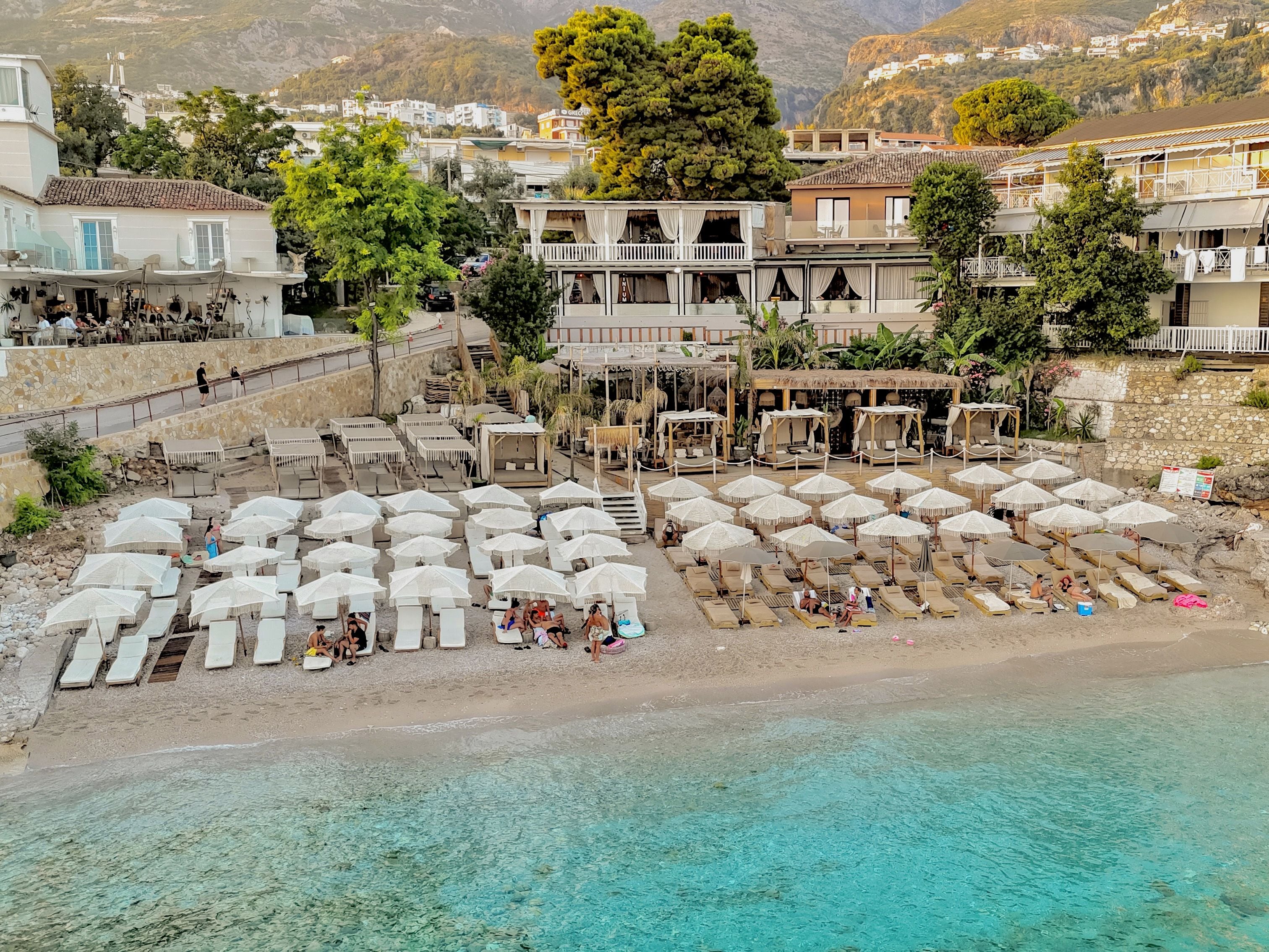 A beachside resort with sun loungers and umbrellas, a pool, and buildings in the background.