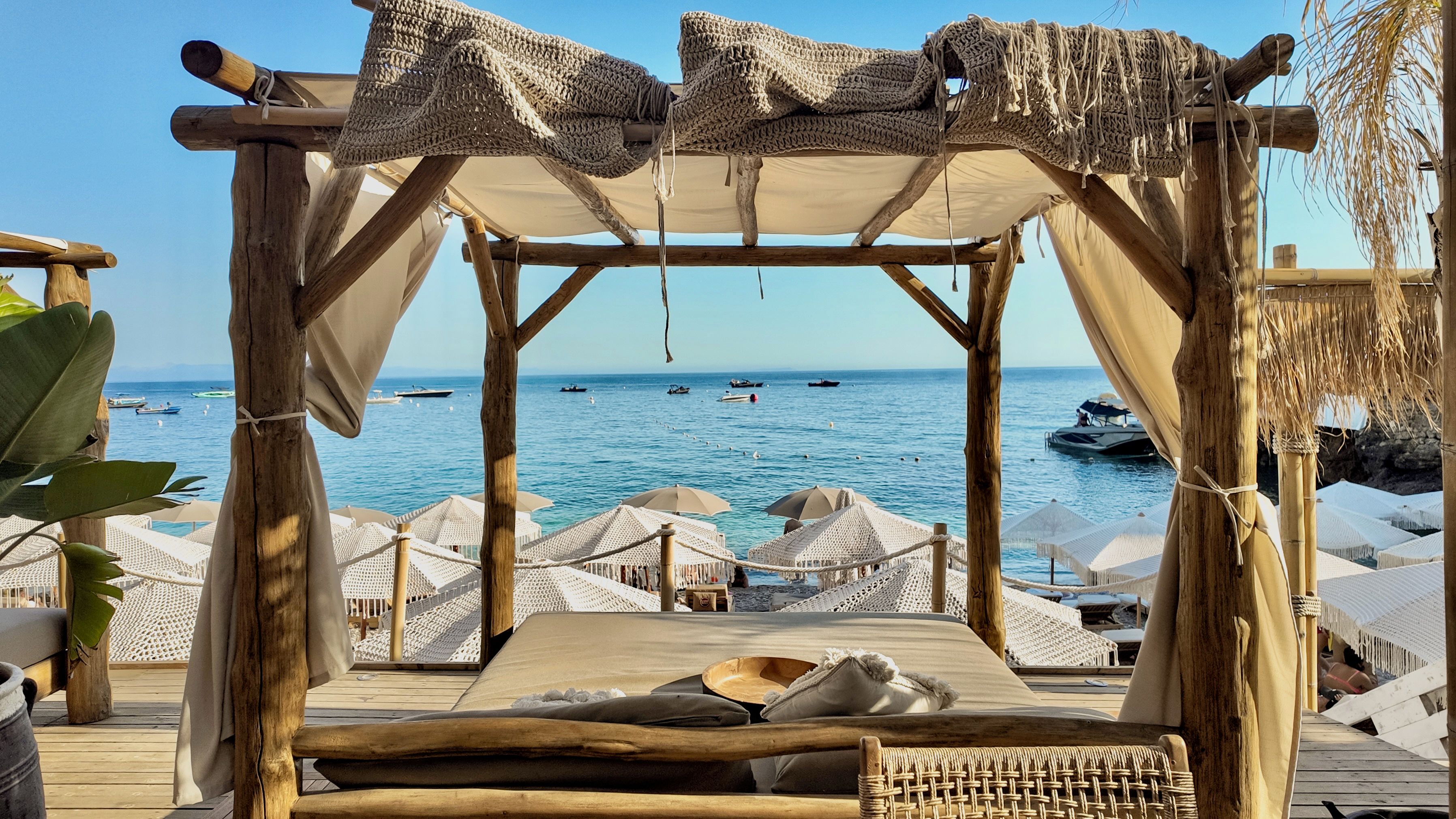 Terraza de madera con toldo que da al mar, con una zona de estar con cojines y vistas a barcos.