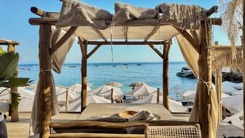 Terraza de madera con toldo que da al mar, con una zona de estar con cojines y vistas a barcos.