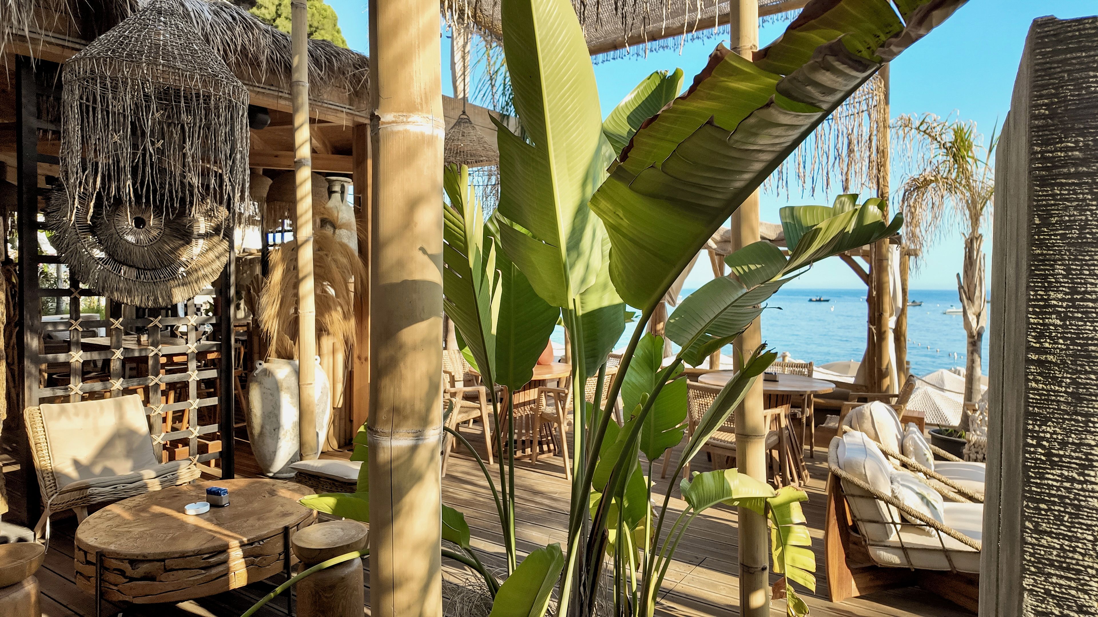 Un espacio al aire libre con mobiliario de madera, techo de paja y vista al mar.