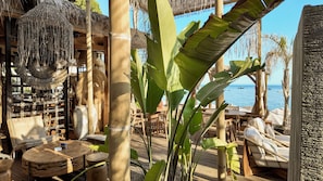 A tropical outdoor seating area with wooden furniture, a thatched roof, and a view of the sea.
