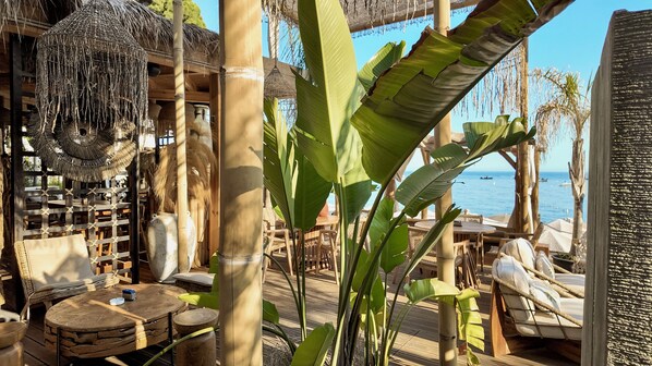 A tropical outdoor seating area with wooden furniture, a thatched roof, and a view of the sea. - Hotel Platinium Dhermi (Dhermi)