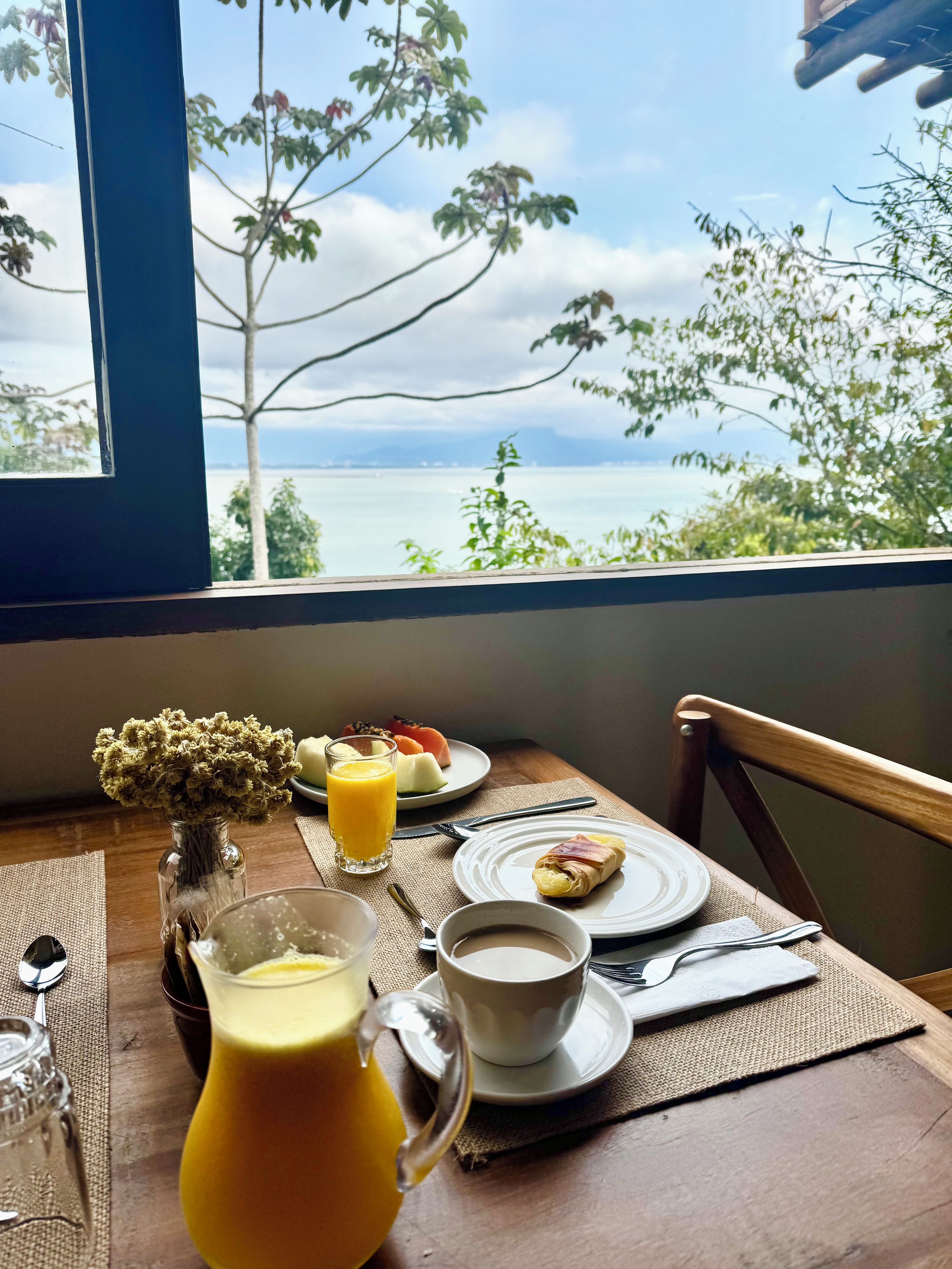 Mesa posta com itens de café da manhã, um copo de suco de laranja e vista para um corpo d'água através de uma janela.
