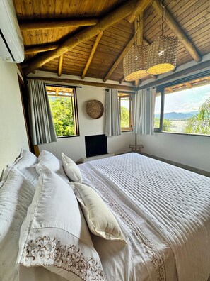 Un dormitorio con una cama grande, vigas de madera en el techo, vista a zonas verdes y una lámpara de fibra trenzada.