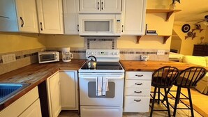 Une cuisine compacte avec des meubles blancs, un four à micro-ondes, une cuisinière et une machine à café. On y trouve deux tabourets de bar et une petite table.
