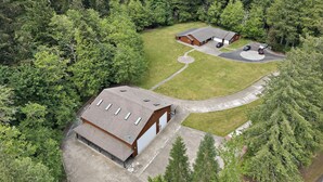 Vista aérea de una propiedad con un edificio grande, una estructura más pequeña adosada y un área de estacionamiento rodeada de bosque denso.