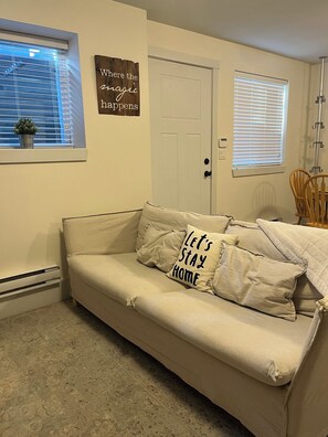 A beige sofa with 'Let's Stay Home' pillows, a white door, a window with blinds, and a small potted plant.