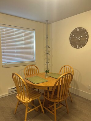 A dining area with a wooden table, four chairs, a clock on the wall, and a window with blinds.