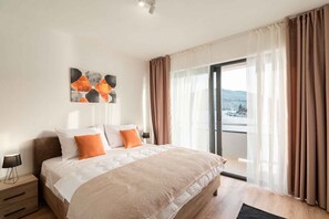 Ein Schlafzimmer mit Bett, Nachttisch, Lampe, einem Fenster mit Vorhängen und Blick ins Freie.