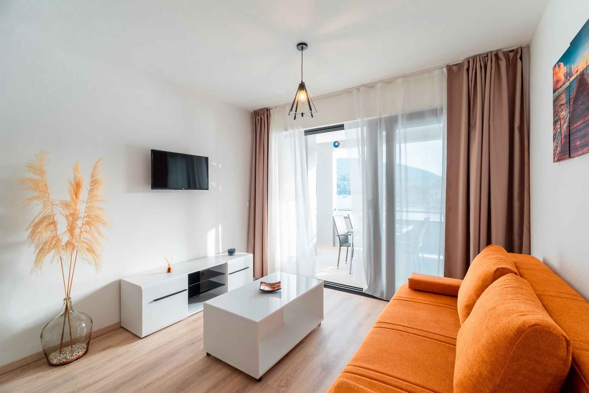 Een moderne woonkamer met een flatscreen-televisie, een witte salontafel, een oranje bank en een groot raam met gordijnen.
