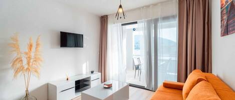 Een moderne woonkamer met een flatscreen-televisie, een witte salontafel, een oranje bank en een groot raam met gordijnen.