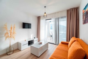 Ein modernes Wohnzimmer mit Flachbildfernseher, weißem Couchtisch, orangefarbenem Sofa und einem großen Fenster mit Vorhängen.