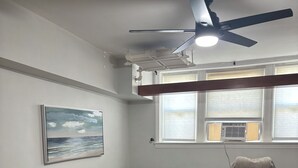Een woonkamer met een hoekbank, een zwarte lederen fauteuil, een klein bijzettafeltje, een plafondventilator en een schilderij aan de muur.