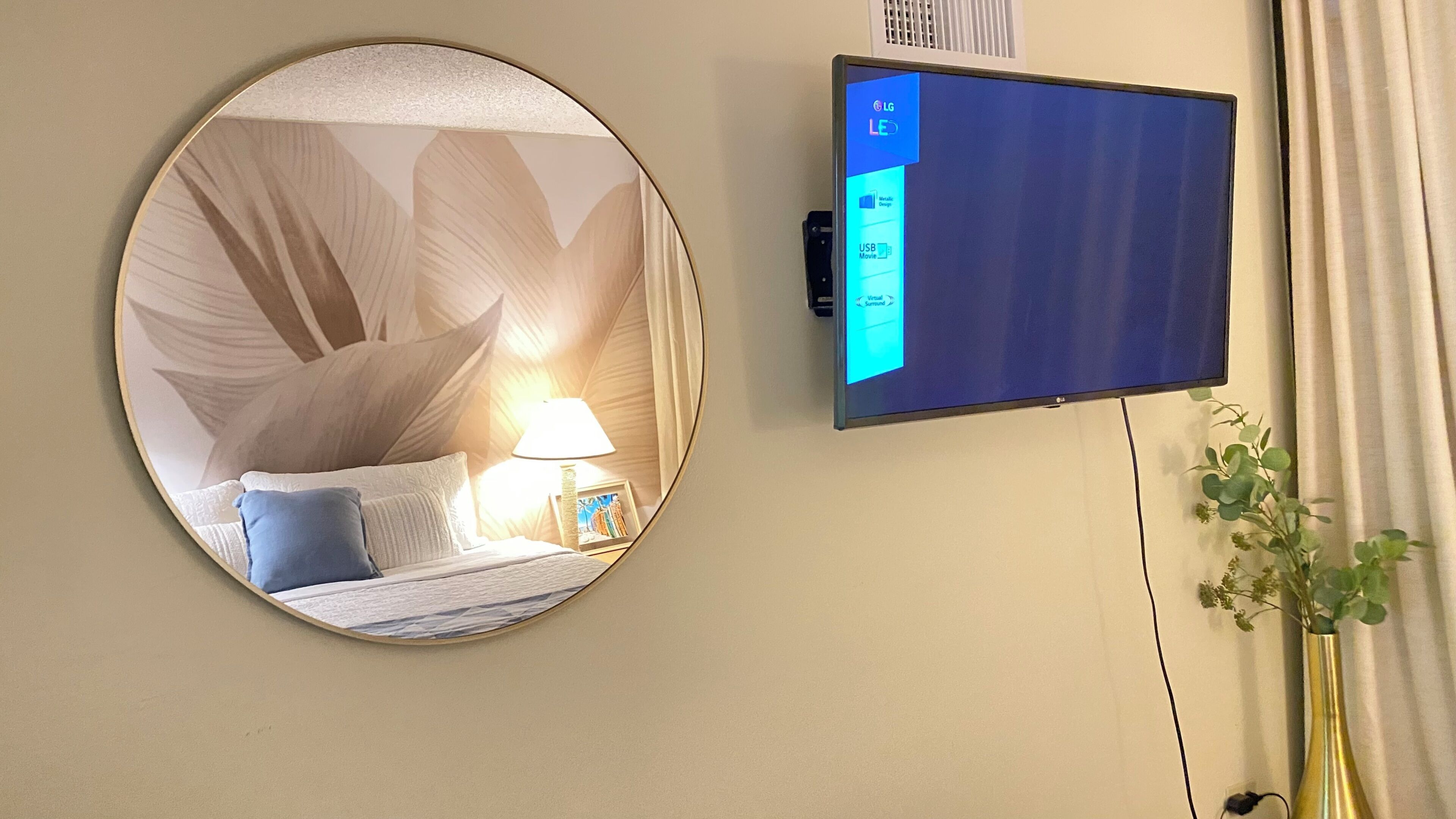 A hotel room with a round mirror, a bed with pillows, a bedside table with a lamp, and a flat-screen TV mounted on the wall.