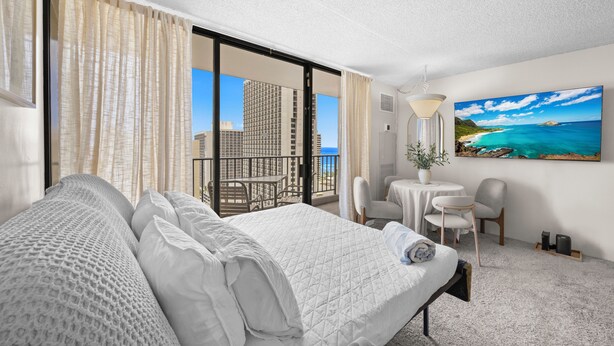 A hotel room with a bed, a balcony with a table and chairs, a painting of a beach, and a view of a cityscape. - Waikiki Cozy &Natural OceanMountain view hotel suite Free-parking  (Honolulu)