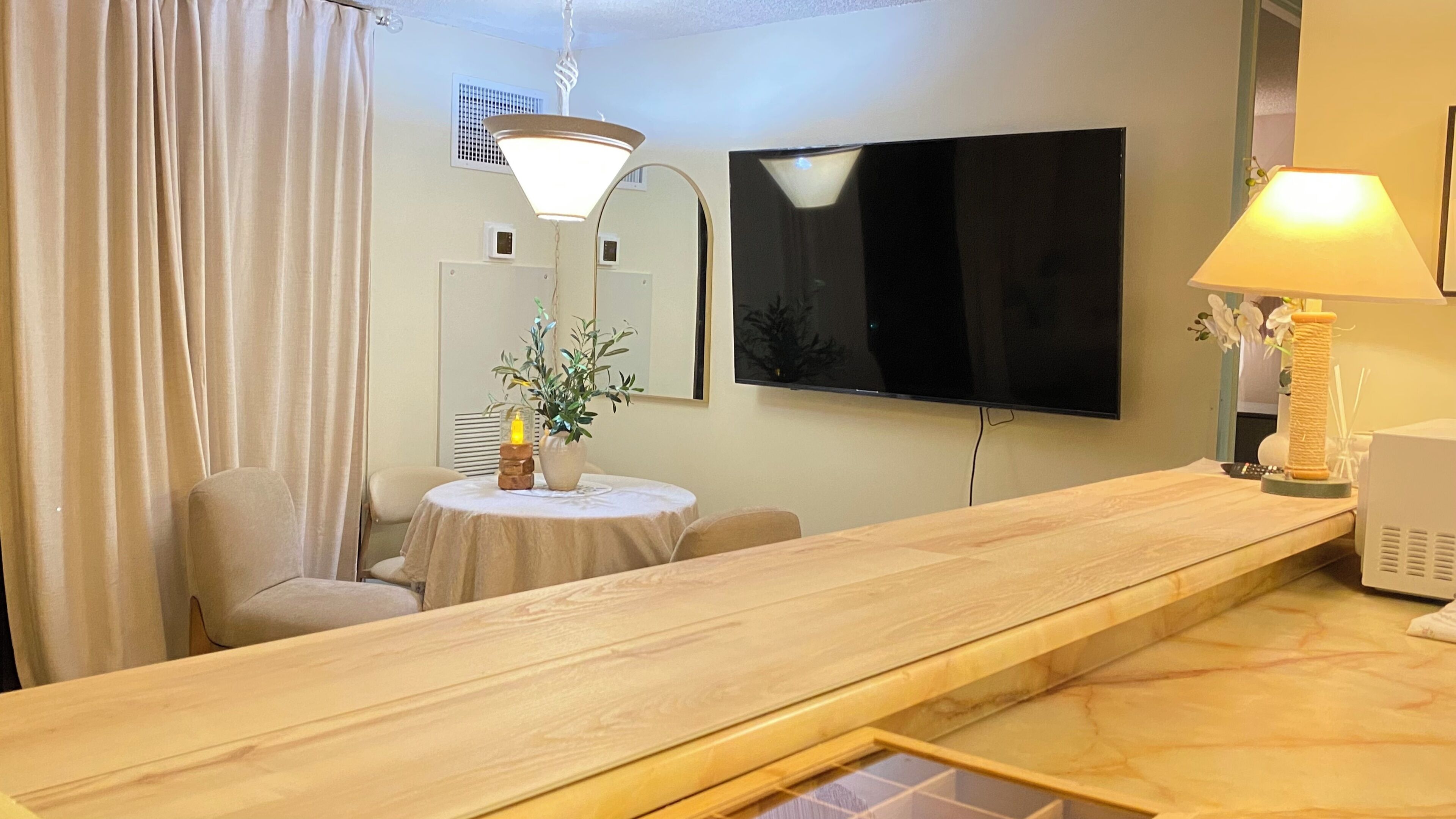 A hotel room with a long wooden table, a round dining table, a TV mounted on the wall, a lamp, and a potted plant.