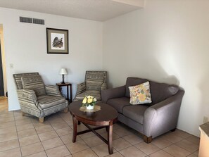 Ein Wohnzimmer mit zwei Sesseln, einem Sofa, einem kleinen runden Tisch mit einer Vase und einem Wandbild.