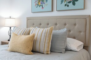 A bed with a tufted headboard, multiple pillows, a bedside table with a lamp, and two framed floral artworks on the wall. - The BurgNest - 2-bedroom townhome in Blacksburg, minutes from Virginia Tech. (Blacksburg)