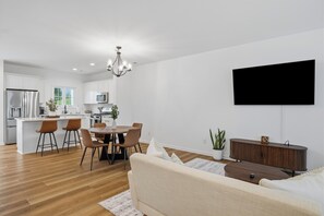 A modern kitchen with white cabinets, a dining area with a round table and chairs, a living room with a sofa and a TV mounted on the wall. - The BurgNest - 2-bedroom townhome in Blacksburg, minutes from Virginia Tech. (Blacksburg)