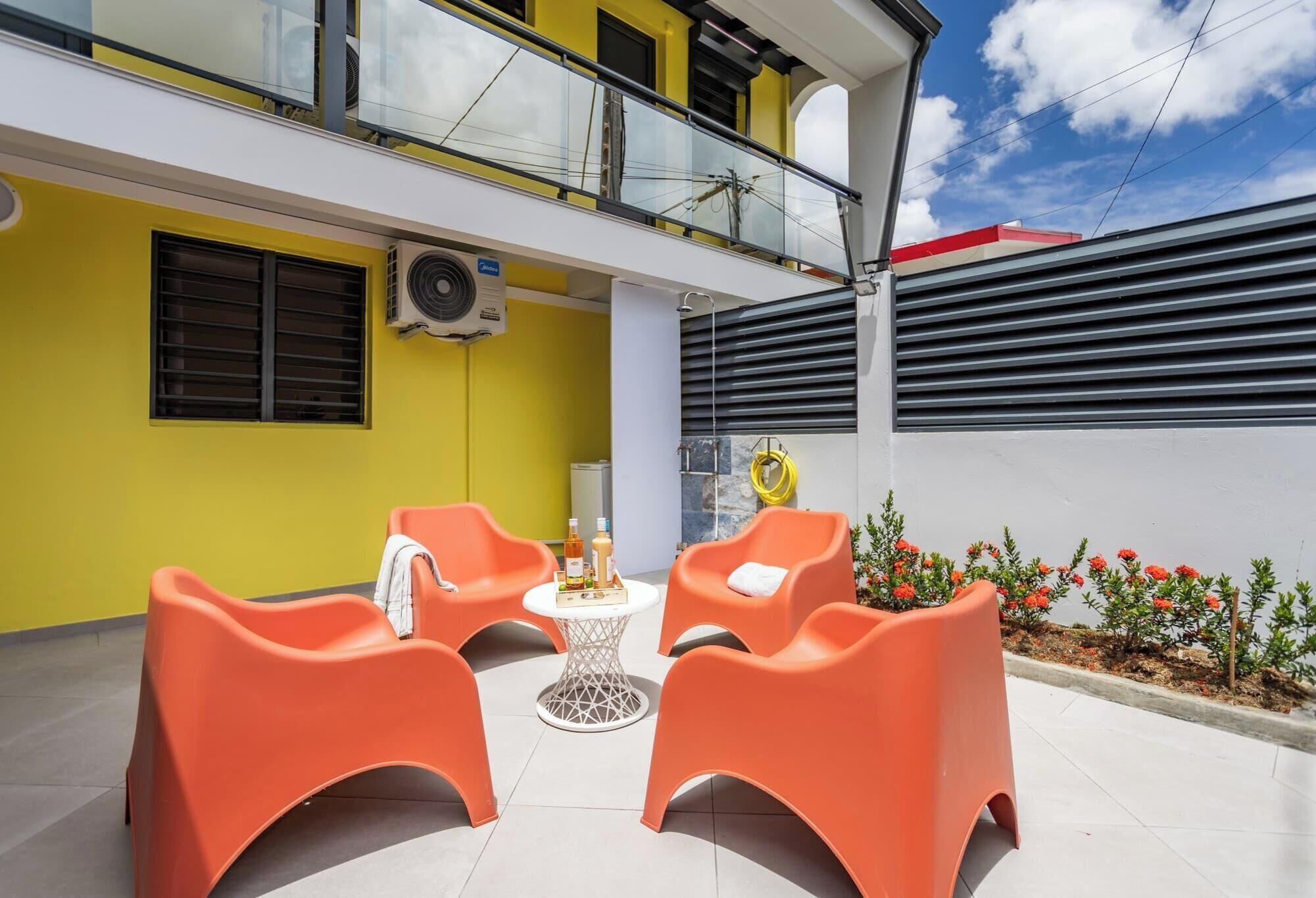 A yellow building with a balcony, orange chairs, a small table, and a flower bed.