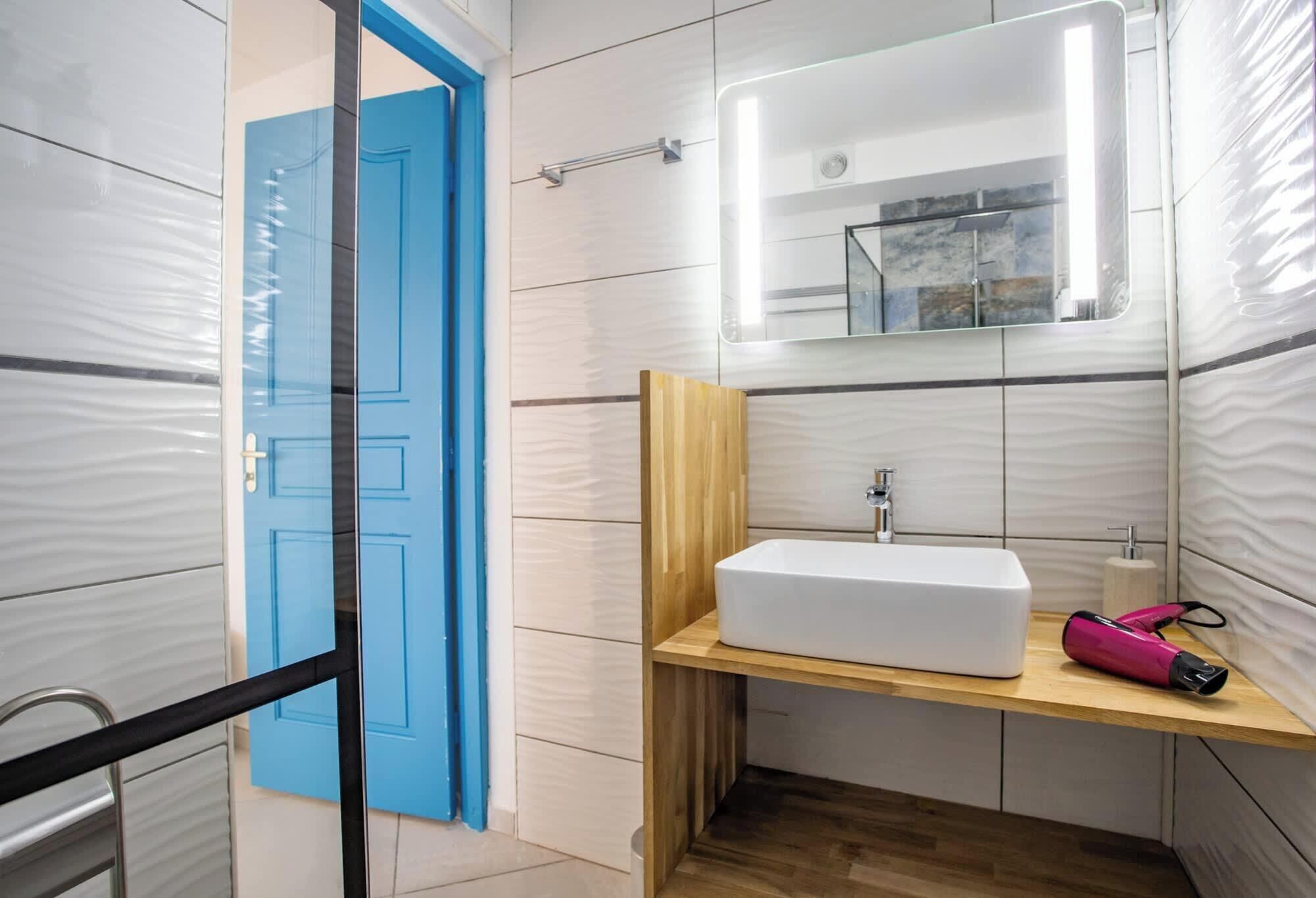 Ein Badezimmer mit blauer Tür, weißem Waschbecken, Spiegel und Holzregal.