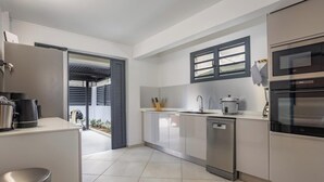 A modern kitchen with a dishwasher, oven, and a window with blinds. - 2BR Apartment Planteur - Beachfront (Les Trois-Îlets)