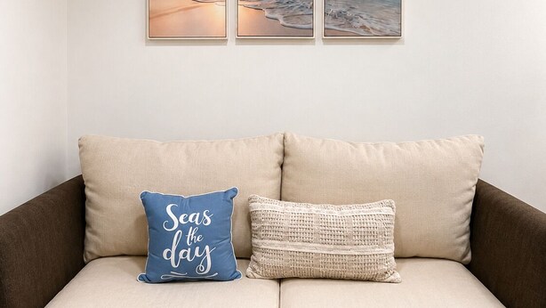 A beige sofa with two pillows, one with text and one textured. Above, three framed beach scenes are hung on the wall. -  Cozy coastal stay in Fort Lauderdale near las olas,Wilton manors,airport,Port (Fort Lauderdale)