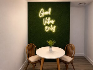 Ein Zimmer mit einer Neonreklame mit dem Schriftzug 'Good Vibes Only' an einer grünen Wand, zwei Holzstühlen, einem kleinen Tisch mit einer Pflanze und einer weißen Deckenleuchte.